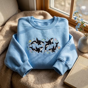 Könnte beinhalten: Ein hellblaues Sweatshirt mit Rundhalsausschnitt und einer Stickerei von drei schwarz-weißen Orcas, umgeben von kleinen bunten Blumen und vierzackigen Sternen. Das langärmelige Kleidungsstück hat einen gerippten Kragen, Bündchen und Saum.