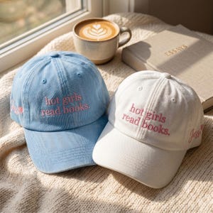 May include: Two baseball caps, one light blue and one white, each featuring the embroidered pink text 'hot girls read books.' on the front and the name 'Jessi' with a small heart icon embroidered on the side panel of each cap.