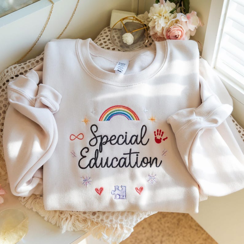 Pode incluir: Camisola creme com as palavras "Special Education" bordadas em escrita preta. Apresenta um arco-&iacute;ris, um s&iacute;mbolo de infinito, uma impress&atilde;o de m&atilde;o, uma pe&ccedil;a de quebra-cabe&ccedil;a, cora&ccedil;&otilde;es e estrelas. A camisola tem mangas compridas.