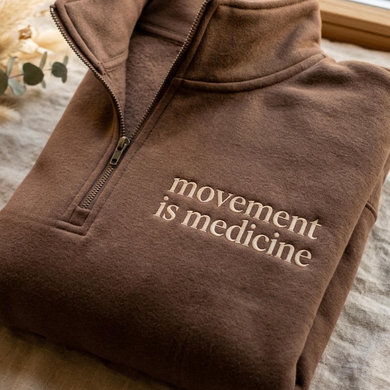 May include: A brown quarter-zip sweatshirt with the embroidered text "movement is medicine." The sweatshirt has a high collar and a silver zipper. The fabric appears soft and comfortable.