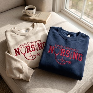 May include: Two crewneck sweatshirts, one cream and one navy blue, each featuring embroidered red text that reads Lewis University NURSING, with a stethoscope forming the letter U and a syringe replacing the letter I in the word nursing.
