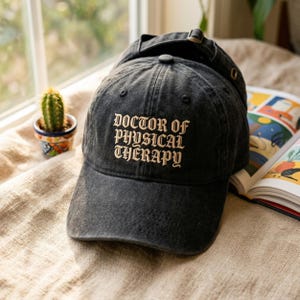 May include: A black baseball cap with the embroidered text "DOCTOR OF PHYSICAL THERAPY" in a decorative font. The cap has a curved brim and an adjustable strap. A small cactus in a decorative pot is visible in the background.