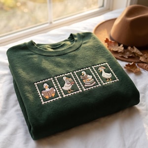 Puede incluir: Una sudadera de cuello redondo verde oscuro con cuatro parches rectangulares bordados, cada uno representando un pato blanco en diferentes posturas con libros. El bordado muestra patos leyendo, cargando y equilibrando pilas de libros.