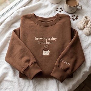 May include: A brown crewneck sweatshirt featuring the embroidered text "brewing a tiny little bean" above a graphic of a smiling coffee cup with a coffee bean. The sleeve is embroidered with the text "est. 2027" in white thread.