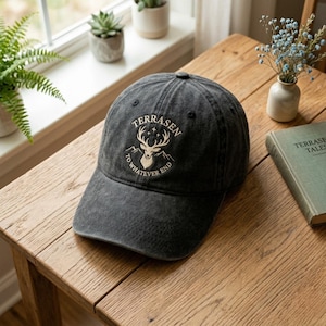 Könnte beinhalten: Eine anthrazitfarbene Baseballkappe mit dem Wort "TERRASEN" über einer Hirschkopf-Grafik und dem Text "TO WHATEVER END" darunter. Die Kappe liegt auf einem Holztisch.
