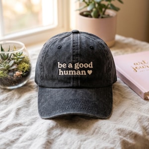 Op de afbeelding: Een antracietgrijze baseballpet met een verwassen, verweerde afwerking. Op de voorkant van de pet staat in wit borduurwerk de tekst "be a good human", gevolgd door een klein wit hartsymbool. De pet heeft een gestructureerde kroon en een gebogen klep.