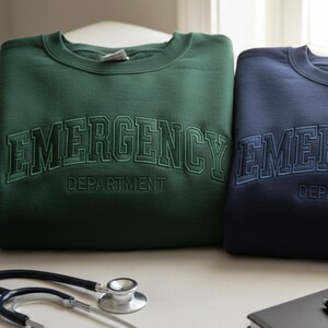 May include: Two sweatshirts, one dark green and one navy blue, with the words "EMERGENCY DEPARTMENT" embroidered across the chest in a collegiate style font. A stethoscope and a notepad are in the foreground.