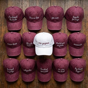 May include: A collection of fourteen baseball caps arranged in a grid. Thirteen caps are a deep maroon colour with white embroidered text, while one central cap is white with maroon text reading mrs. guzman. The other caps feature various phrases.