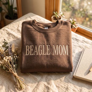 Könnte beinhalten: Ein dunkelbraunes Sweatshirt mit Rundhalsausschnitt und dem Text BEAGLE MOM, der in einer hellbeigen Serifenschrift auf die Brust gestickt ist. Das Kleidungsstück hat einen gerippten Kragen und lange Ärmel und liegt flach gefaltet.