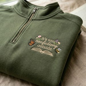 May include: Olive green quarter-zip sweatshirt with embroidered design. The design includes a butterfly, flowers, a bee, and an open book with the text "it's too nonfiction outside". The sweatshirt has a zipper closure and a high collar.