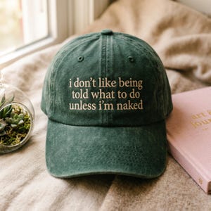 Könnte beinhalten: Dunkelgrüne Baseballkappe mit dem gestickten Text "i don't like being told what to do unless I'm naked". Der Hut ist aus weichem Stoff und hat einen gebogenen Schirm. Im Hintergrund ist ein rosa Buch zu sehen.