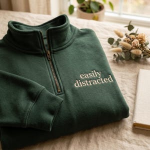 May include: A dark green sweatshirt with a quarter-zip neckline and the embroidered text "easily distracted" in white. The sweatshirt is folded on a light-colored surface.