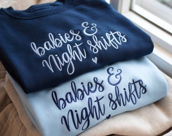 May include: Two sweatshirts, one navy blue and one light blue, are stacked on top of each other. The navy blue sweatshirt has white embroidered text that reads "babies & Night shifts" with a small heart. The light blue sweatshirt has the same text in navy blue.