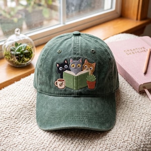 May include: A forest green baseball cap featuring an embroidered design of three cats reading a green book. The embroidery includes a black cat, a grey cat, and a tabby cat, accompanied by a small coffee mug and a potted succulent plant.