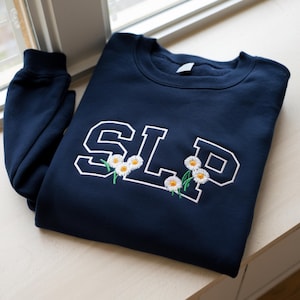 Può includere: Felpa blu navy con le lettere "SLP" ricamate in bianco, decorate con piccoli fiori di margherita. La felpa è piegata ordinatamente, con una manica visibile. Su una superficie chiara.