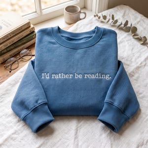 May include: A blue crewneck sweatshirt featuring the white embroidered text "I'd rather be reading." centred on the chest. The garment has long sleeves with ribbed cuffs and a ribbed neckline, constructed from a solid-coloured fabric material.