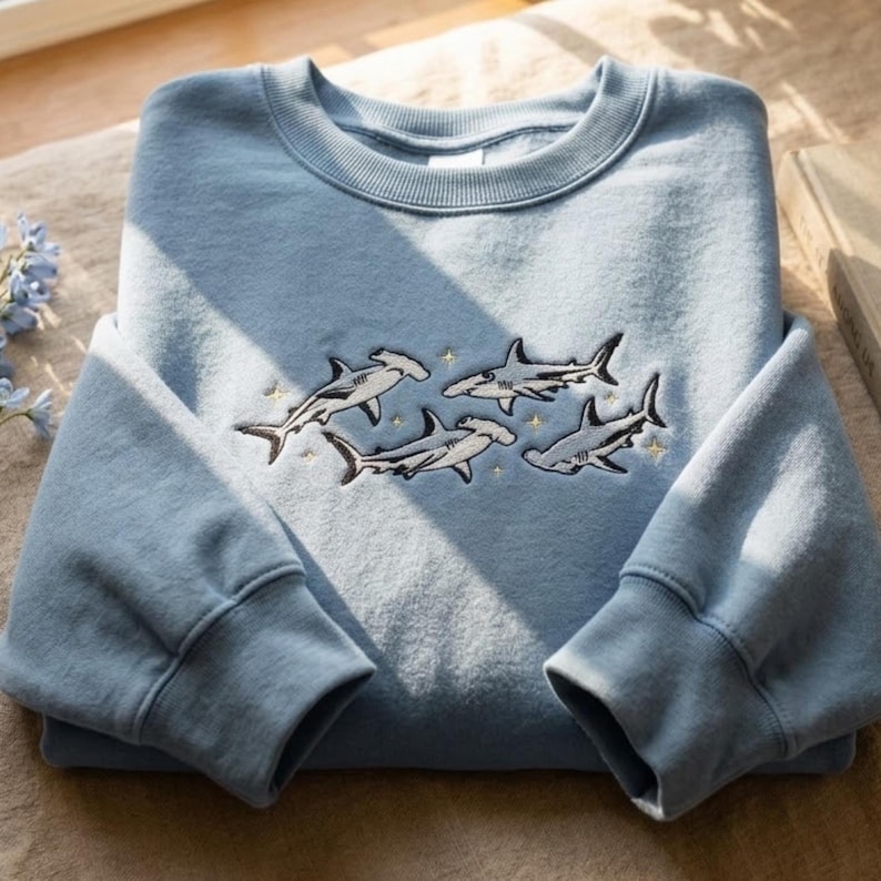 May include: A light blue crewneck sweatshirt featuring an embroidered design of four hammerhead sharks swimming among small yellow stars. The garment has long sleeves with ribbed cuffs and a ribbed neckline, showcasing a detailed marine theme.