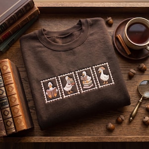 May include: A dark brown crewneck sweatshirt featuring an embroidered design of four rectangular postage stamp frames. Each frame contains a white duck interacting with books, including reading, carrying a stack, sitting on books, and wearing one.