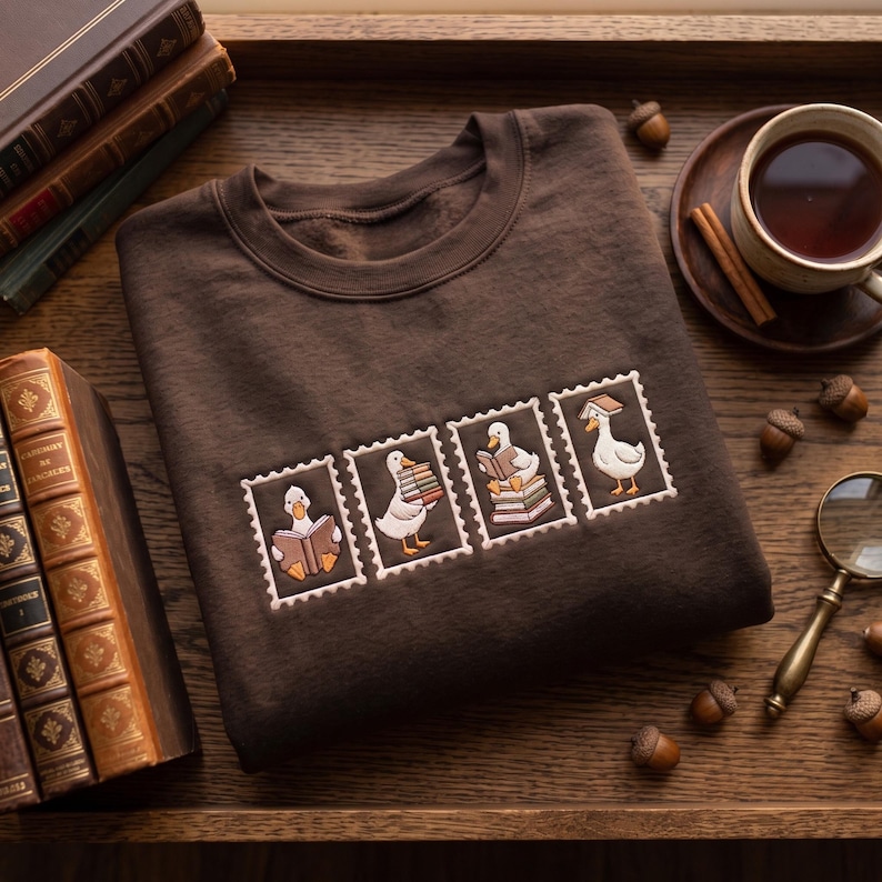 May include: A dark brown crewneck sweatshirt featuring an embroidered design of four rectangular postage stamp frames. Each frame contains a white duck interacting with books, including reading, carrying a stack, sitting on books, and wearing one.