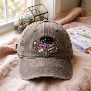 May include: A brown, distressed-finish baseball cap featuring an embroidered design of a black cat sleeping on a stack of two books, one purple and one brown, accented with small white and purple flowers and tiny star-like details.