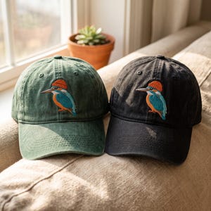 Peut inclure: Deux casquettes de baseball, une vert foncé et une noire, arborant chacune une broderie représentant un martin-pêcheur bleu et orange portant un petit bonnet en tricot orange. Les casquettes ont une calotte structurée et une visière courbée.
