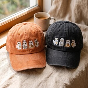 May include: Two baseball caps, one in a faded orange and one in a faded black, each featuring an embroidered design of four white cats engaged in baking activities, including holding a jar, mixing in a bowl, slicing bread, and holding a loaf.