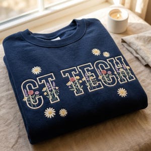 May include: Navy blue sweatshirt with the embroidered text "CT TECH" in a white outline, filled with colorful floral designs. Additional embroidered daisies are scattered around the text. The sweatshirt is folded neatly on a neutral surface.
