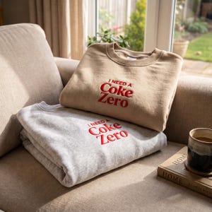 Op de afbeelding: Twee sweaters met ronde hals, één in beige en één in grijs gemêleerd, elk voorzien van de geborduurde rode tekst "I NEED A Coke Zero" in het midden op de borst. De kledingstukken hebben geribbelde kragen en boorden en een eenvoudig ontwerp met lange mouwen.