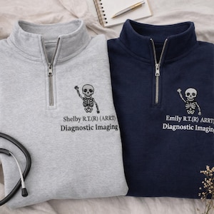 Può includere: Due felpe con zip a un quarto, una grigia e una blu navy. Ognuna presenta una grafica a scheletro e il testo "Shelby R.T.(R) (ARRT) Diagnostic Imaging" e "Emily R.T.(R) ARRT Diagnostic Imaging". È visibile uno stetoscopio.