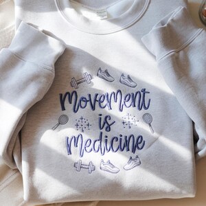 May include: A light gray sweatshirt with the embroidered text "Movement is Medicine" in dark blue script. The design includes illustrations of a barbell, sneakers, and tennis rackets, promoting a fitness theme.