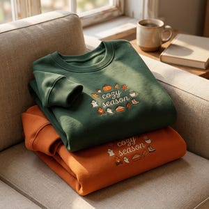 May include: A dark green sweatshirt and an orange sweatshirt stacked on a sofa. Both feature embroidered text that reads cozy season, surrounded by small icons of a pumpkin, leaves, a mug, boots, a pie, a campfire, and ghosts.