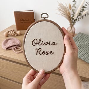 May include: An oval wooden embroidery hoop frame containing a cream-colored fabric insert. The name Olivia Rose is displayed in dark brown, raised script lettering centered on the fabric. A small bronze-toned metal hanging loop is attached to the top.