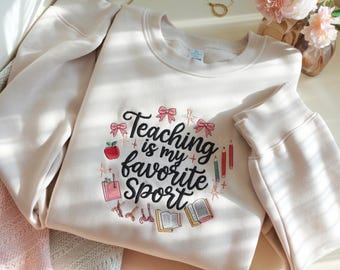 May include: A beige sweatshirt with the phrase "Teaching is my favorite Sport" embroidered in black script with colorful school-themed graphics. The design includes bows, an apple, pencils, books, and a school bag. The sweatshirt has a crew neck and long sleeves.