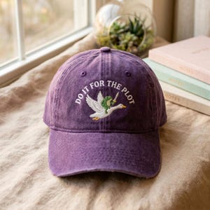 May include: A purple baseball cap with a distressed finish featuring an embroidered graphic of a green frog holding a sword while riding a white goose. The text "DO IT FOR THE PLOT" is arched in white embroidery above the central animal illustration.