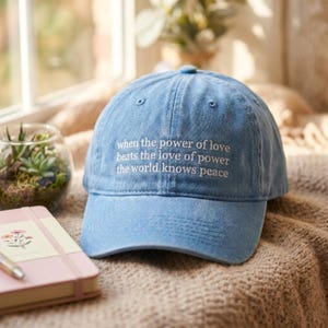 May include: A light blue baseball cap with a distressed finish featuring white embroidered text that reads: when the power of love beats the love of power the world knows peace. The cap has a structured crown and a curved brim with stitching.