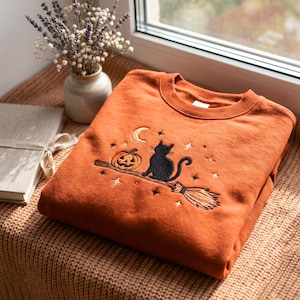 Puede incluir: Una sudadera de cuello redondo color naranja quemado con un bordado de un gato negro sentado sobre una escoba junto a una calabaza. El diseño incluye una luna creciente y pequeñas estrellas cosidas con hilo blanco y dorado.