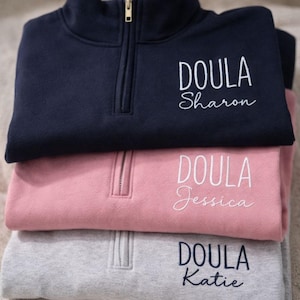 May include: Three stacked quarter-zip pullovers in navy, pink, and grey. Each features the word "DOULA" embroidered in white, with a name below. The navy pullover says "Sharon", the pink says "Jessica", and the grey says "Katie".