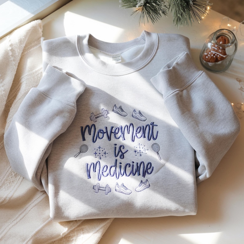 May include: A light gray sweatshirt with the text "Movement is Medicine" in blue script. The design includes illustrations of dumbbells, sneakers, and tennis rackets. The sweatshirt is displayed on a white surface.