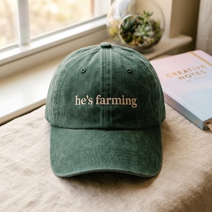 Może przedstawiać: Ciemnozielona czapka bejsbolówka z haftowanym napisem "he's farming" w jasnobeżowym kolorze. Czapka ma zakrzywiony daszek i klasyczny sześciopanelowy design. Kapelusz jest wykonany z miękkiej, teksturowanej tkaniny.