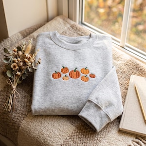 Pode incluir: Uma camisola de gola redonda cinzenta clara com um design bordado de sete abóboras cor de laranja de vários tamanhos dispostas numa fila horizontal no centro do peito. O tecido tem uma textura mesclada e mangas compridas com punhos.