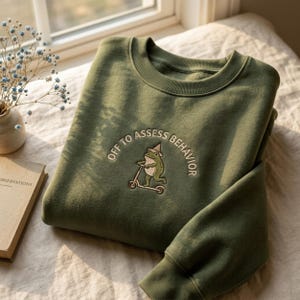 Könnte beinhalten: Olivgrünes Sweatshirt mit der gestickten Aufschrift "OFF TO ASSESS BEHAVIOR" um einen Cartoon-Frosch auf einem Roller. Das Sweatshirt hat einen Rundhalsausschnitt und gerippte Bündchen und Saum.