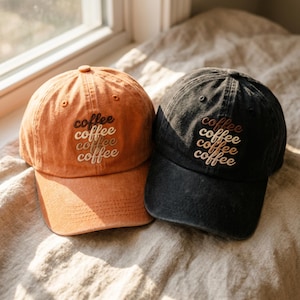 May include: Two baseball caps, one in a washed orange and one in a washed black, each featuring the word coffee embroidered four times in stacked, varying shades of brown and cream thread across the front panels of the six-panel crown.
