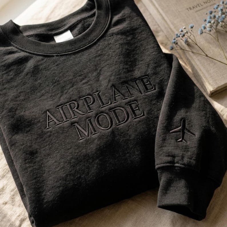 May include: Black sweatshirt with the words "AIRPLANE MODE" embroidered across the chest. A small airplane graphic is embroidered on the left sleeve. The sweatshirt is folded on a neutral surface.