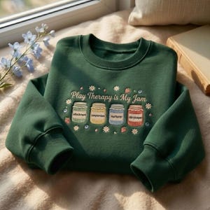 Peut inclure: Un sweat-shirt à col rond vert foncé avec une broderie indiquant Play Therapy is My Jam au-dessus de quatre bocaux colorés étiquetés attachment, compromises, nurturer et storykeeper, entourés de petites fleurs, baies et feuilles.