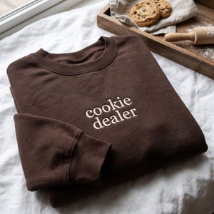 May include: A dark brown crewneck sweatshirt featuring the text cookie dealer embroidered in a light cream-colored thread across the center of the chest. The garment has long sleeves with ribbed cuffs and a ribbed neckline, made of a solid fabric.
