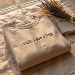 May include: A beige crewneck sweatshirt featuring the embroidered text "wait, i see a bug" in a dark brown thread. The garment has a ribbed collar and long sleeves, presented in a folded position with a neutral tone and a soft texture.