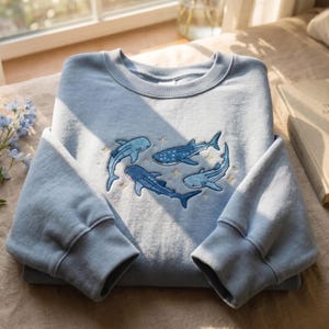 Op de afbeelding: Een lichtblauw sweatshirt met ronde hals en een geborduurd ontwerp van drie walvishaaien die in een cirkelvormige formatie zwemmen, geaccentueerd met kleine gouden sterdetails. Het kledingstuk heeft lange mouwen met geribbelde boorden.