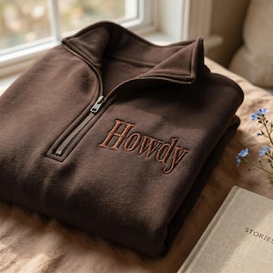 May include: A dark brown quarter-zip sweatshirt featuring a high collar and a metal zip. The word Howdy is embroidered in a matching dark brown thread on the upper left chest area of the garment, which is folded neatly.