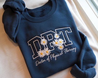 May include: A navy blue sweatshirt featuring the letters "DPT" in white with a navy outline, decorated with daisy motifs. The words "Doctor of Physical Therapy" are embroidered below. The garment is folded on a white surface, near a window.