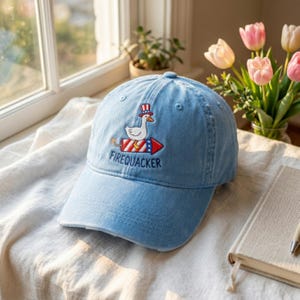 May include: Light blue baseball cap with the embroidered word "FIREQUACKER" and a cartoon duck wearing a top hat and riding a firework. The cap is made of a soft, faded fabric.